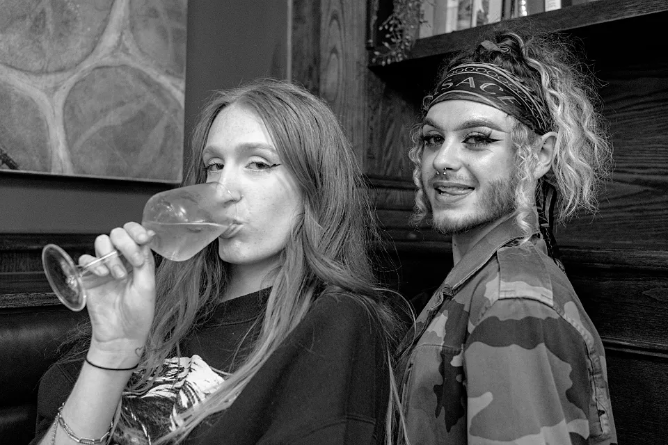 In the corner of a bar in Elephant and Castle, 2 friends pose for the camera: one drinks expressionlessly the other runs their tongue across their teeth