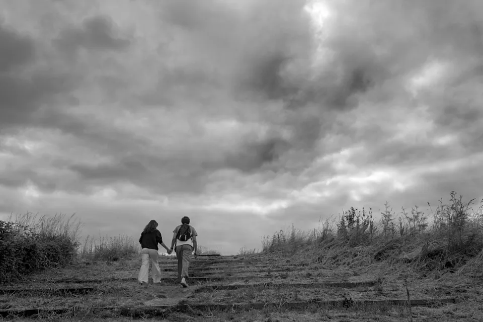 Young couple, hand-in-hand, climb a grassy incline towards a cloudy sky.