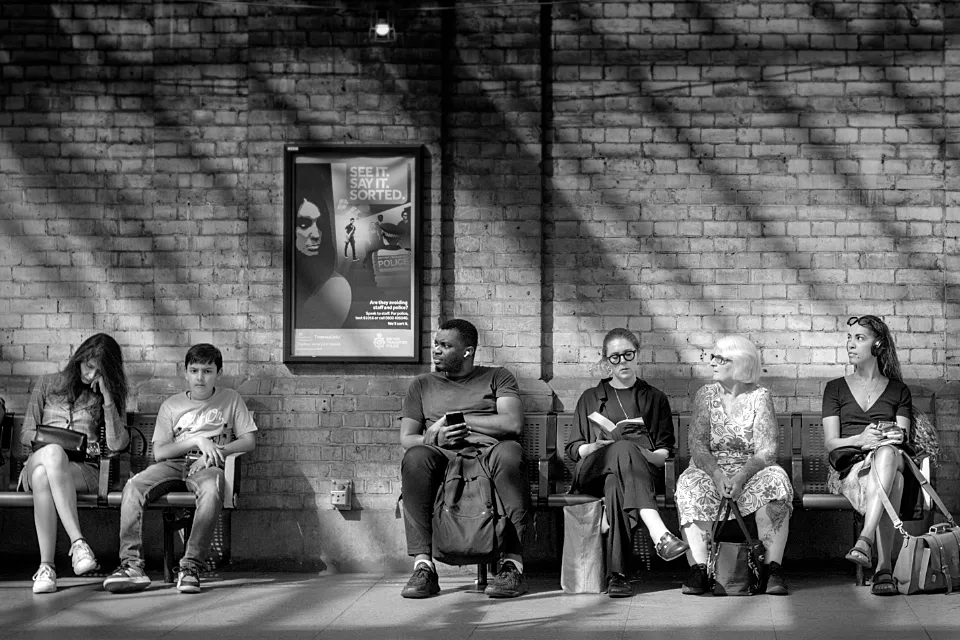 Diagonal streaks of light fall on a brick wall behind passengers who sit waiting on a railway platform in Farringdon Station 