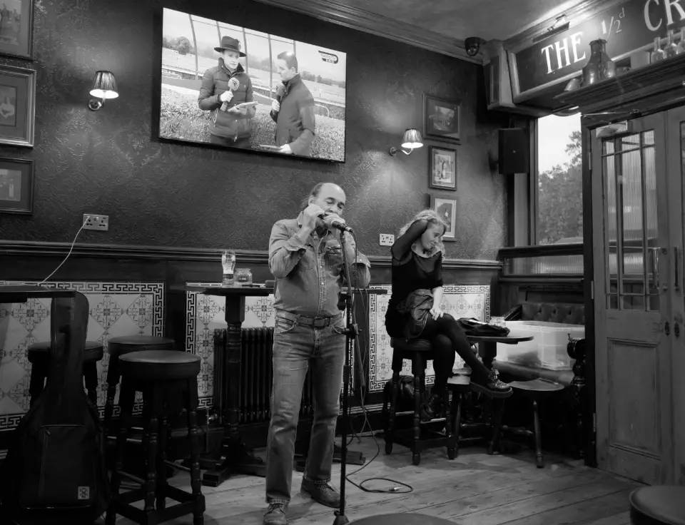 In a traditional pub interior, a bald man concentrates on playing his mouth organ into a microphone while the event host appears a little bored in the background