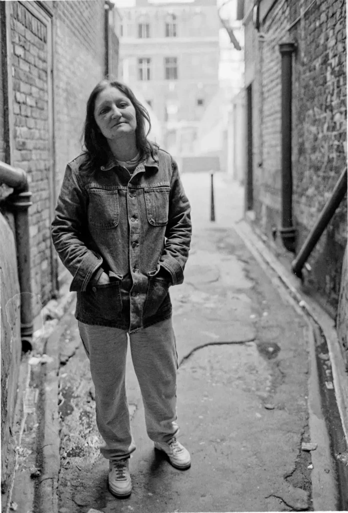 Full-length photo of a beaten woman standing with her hands in her pockets in a Soho alleyway. She looks challengingly towards the camera